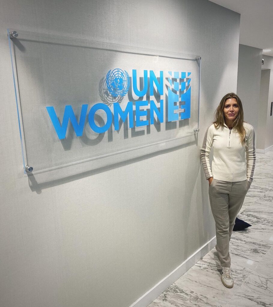 A woman stands next to a wall sign that reads UN Women in blue letters in a modern, marble-floored hallway.