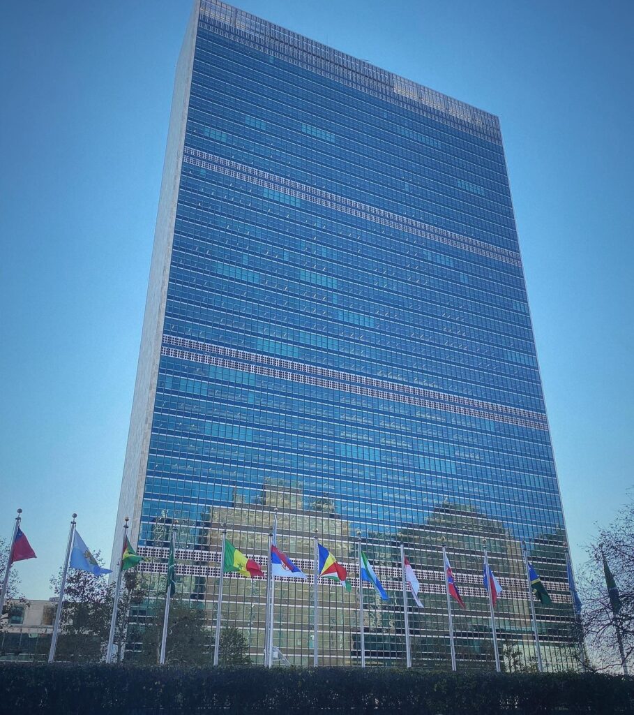 A tall glass office building with multiple national flags displayed on poles in front, set against a clear blue sky.