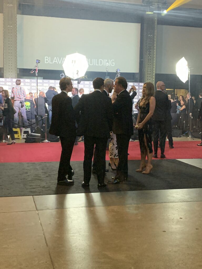A group of people dressed in formal attire stand and talk on a red carpet at an indoor event, with photographers and bright lights in the background.
