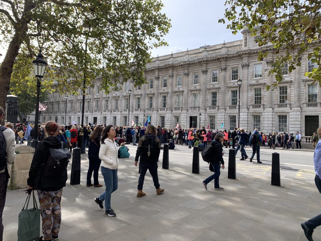 A crowd of people gathers on a city sidewalk and street near a large, historic building with columns and many windows on a cloudy day.