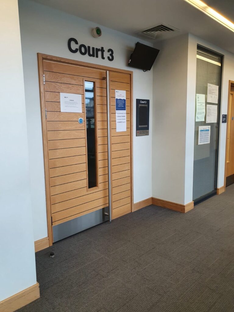 A closed wooden door labeled Court 3 in a hallway, with notices posted on the door and walls, and a television screen above the door.