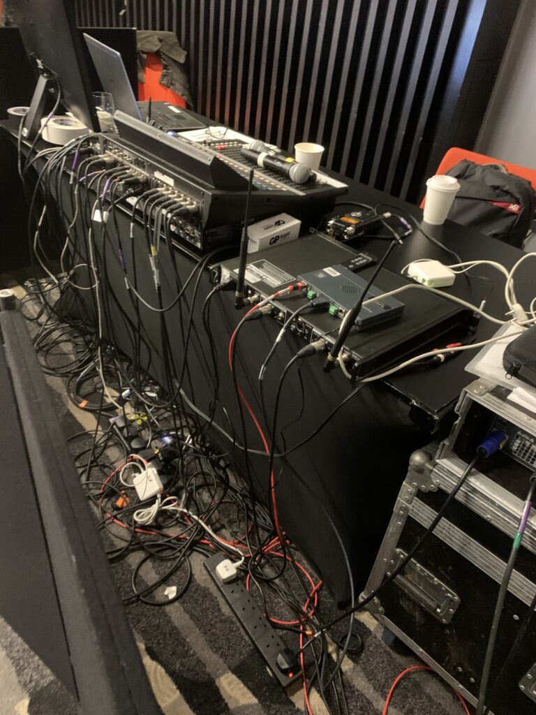 Audio-visual equipment setup with numerous cables, laptops, and electronic devices on tables, indicating a technical control area at an event.