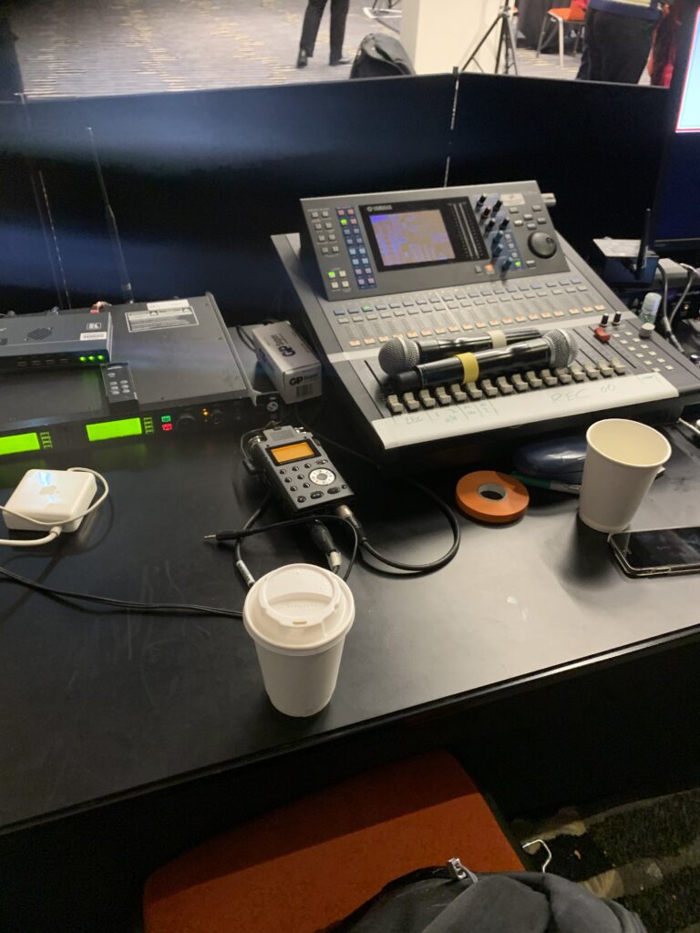 Audio-visual control desk with a sound mixer, recording devices, cables, two paper cups, a smartphone, and other equipment on a black table.