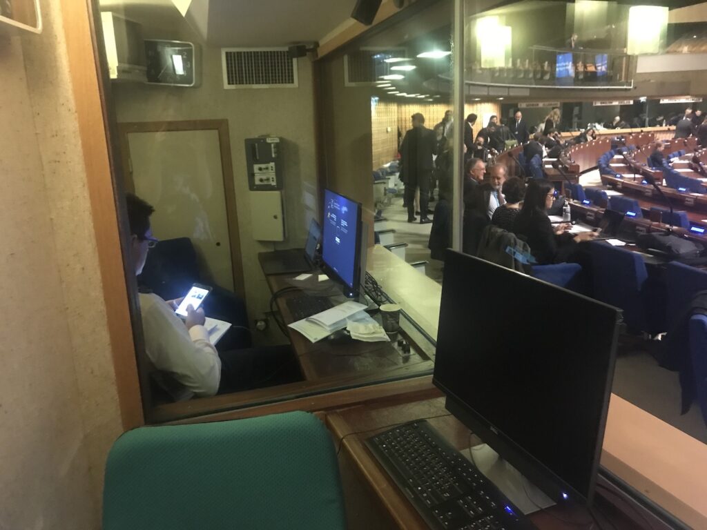 A person sits in a small booth with computer monitors, looking at a phone, while a conference or meeting takes place in the adjacent room visible through glass.