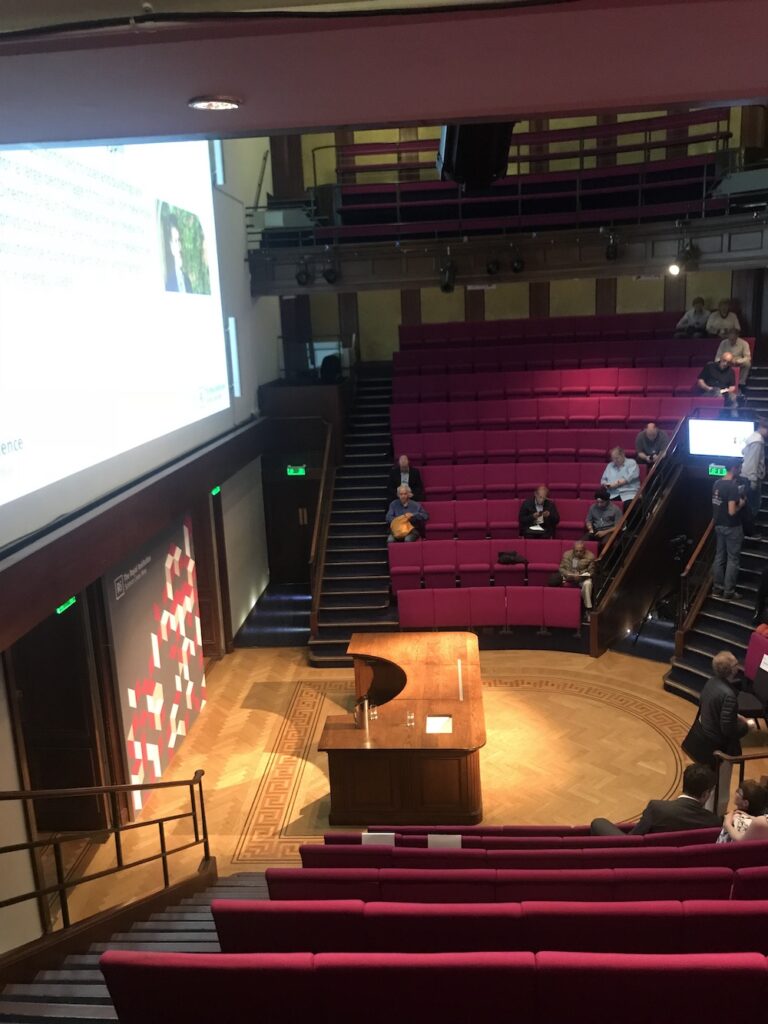 An empty lecture hall with pink tiered seating, a large projection screen displaying a presentation, and a few people seated sparsely around the room.